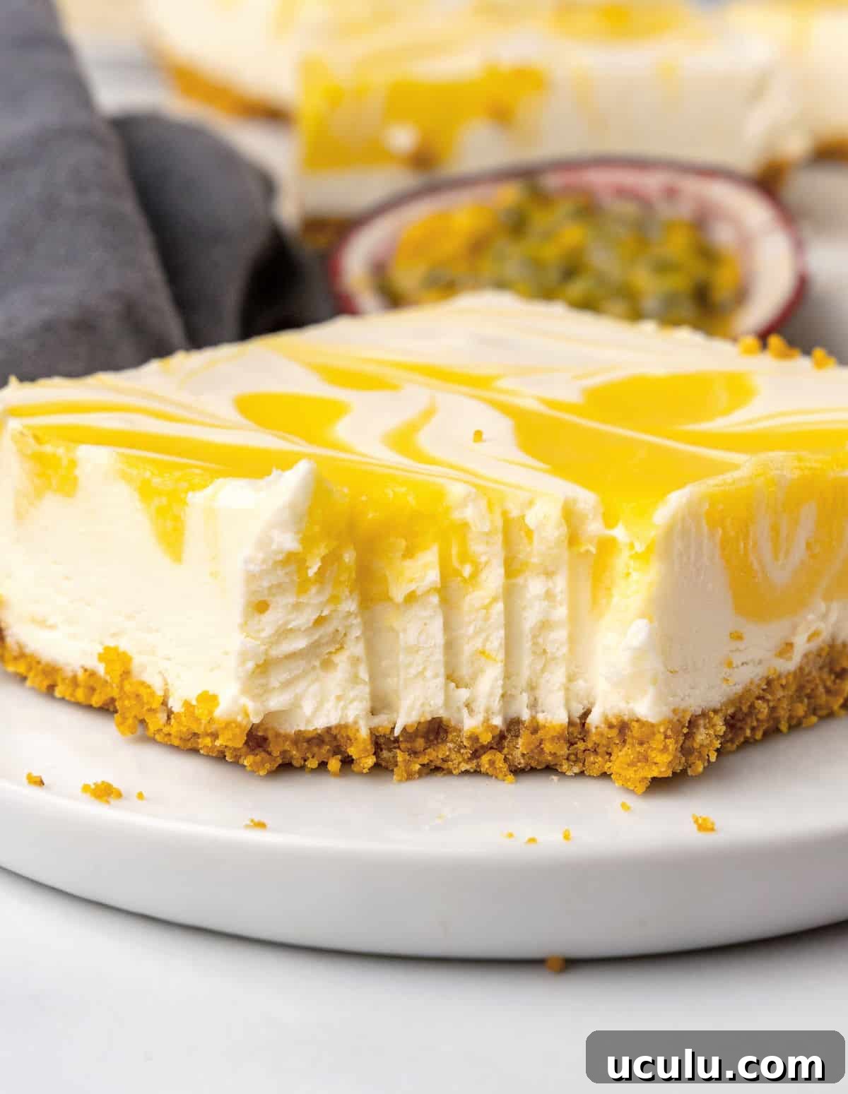 Bitten square of passion fruit no bake cheesecake on a white plate. Blue cloth and fresh passionfruit in the background. 