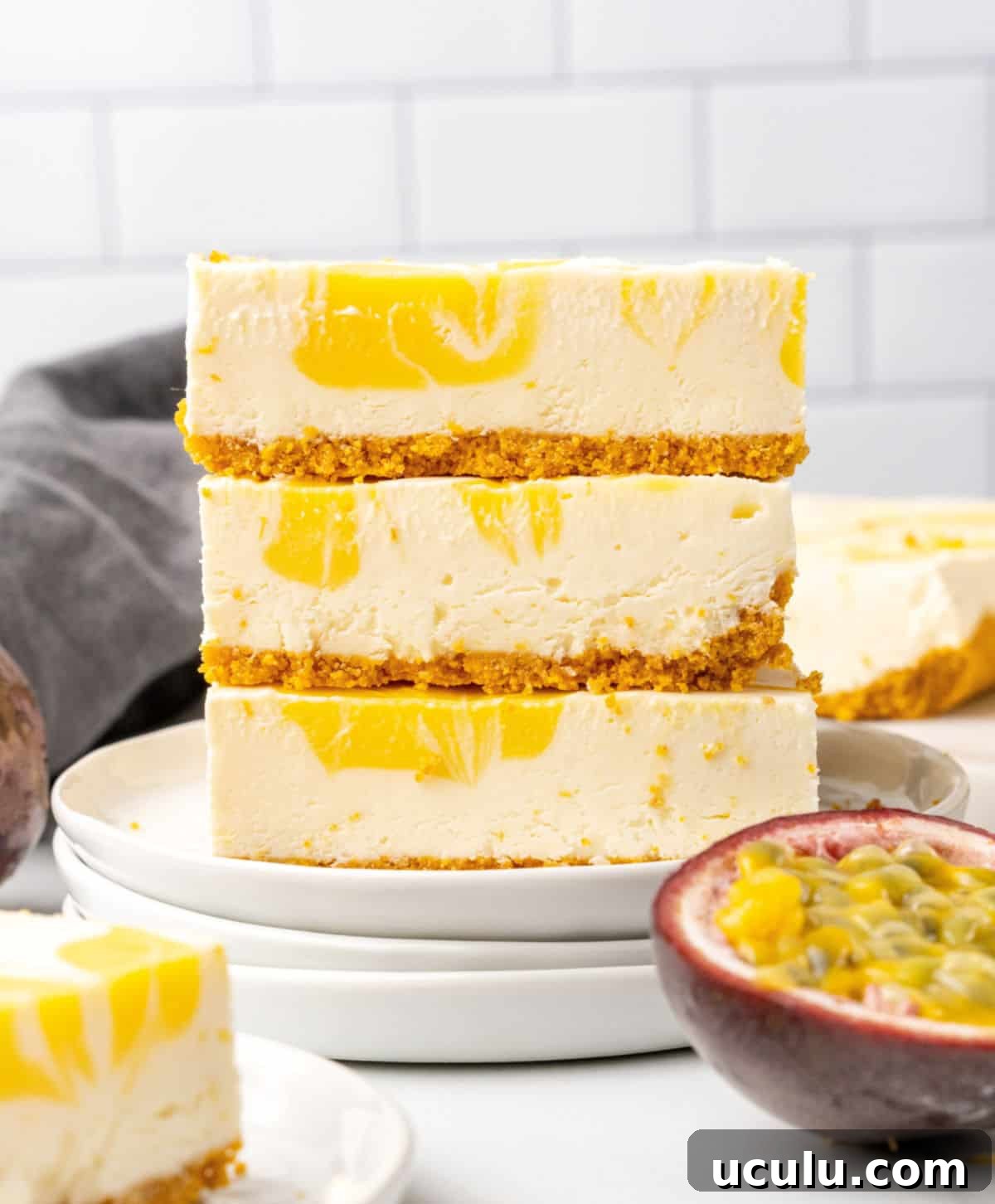 On a stack of white plates, three squares of cheesecake with passion fruit are stacked. White tile background, fresh half fruit, blue cloth. 