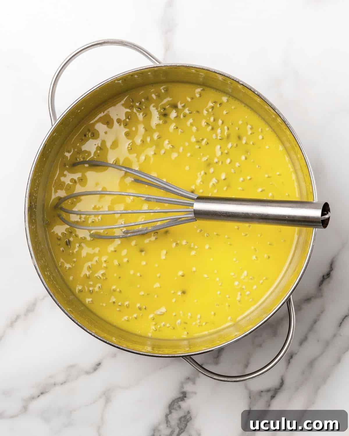 Metal saucepan and whisk with passion fruit curd on white marble.