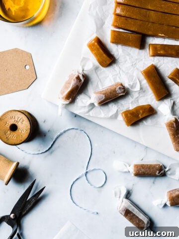 Pieces of candy being cut into individual pieces and wrapped. The work surface is grey marble. There are brown paper gift tags, a vintage spool of twine, and scissors nearby. A glass of whiskey peeks into the frame at top left.