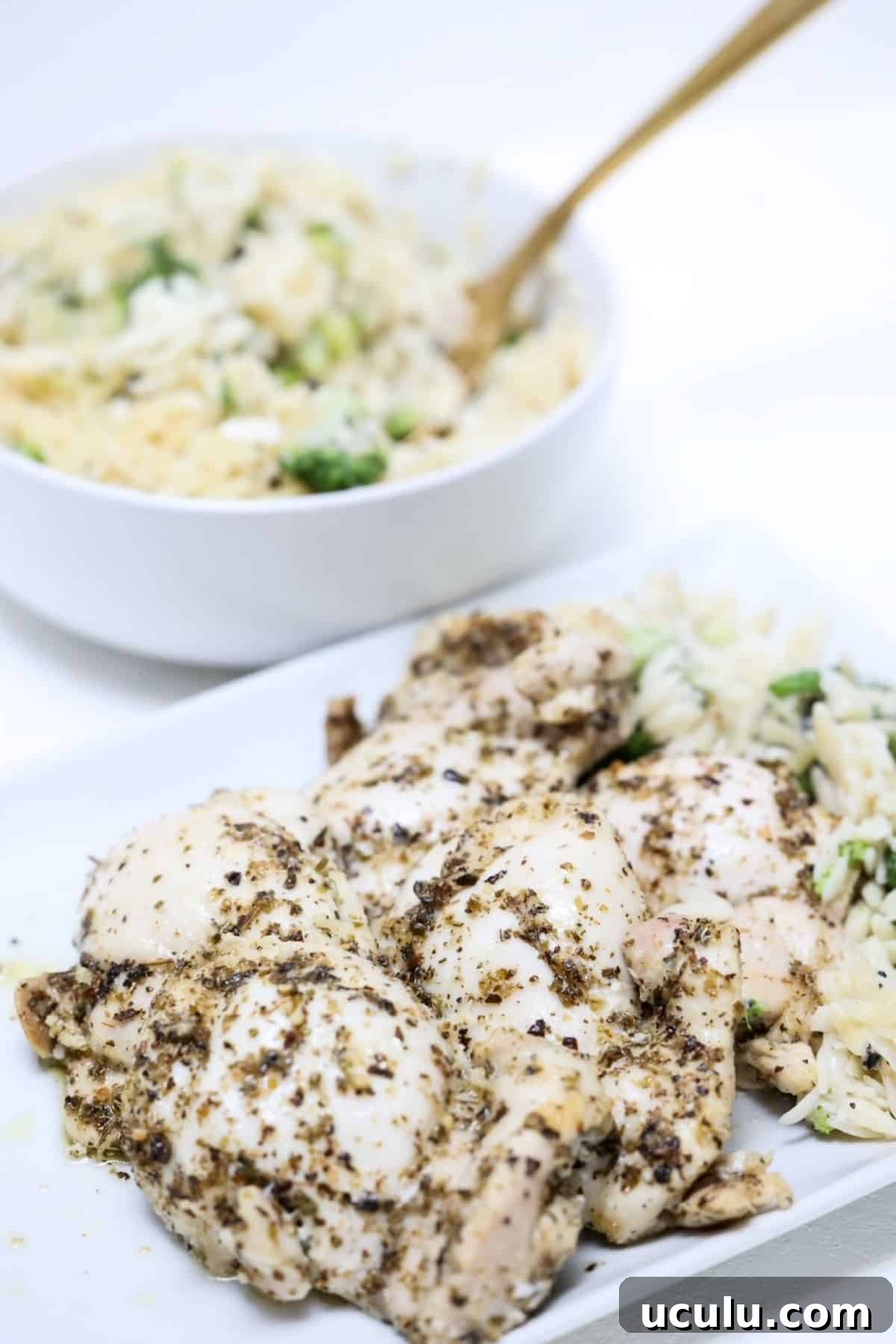 Pieces of baked Italian chicken thighs on a white plate with a bowl of orzo in a white bowl behind the plate.