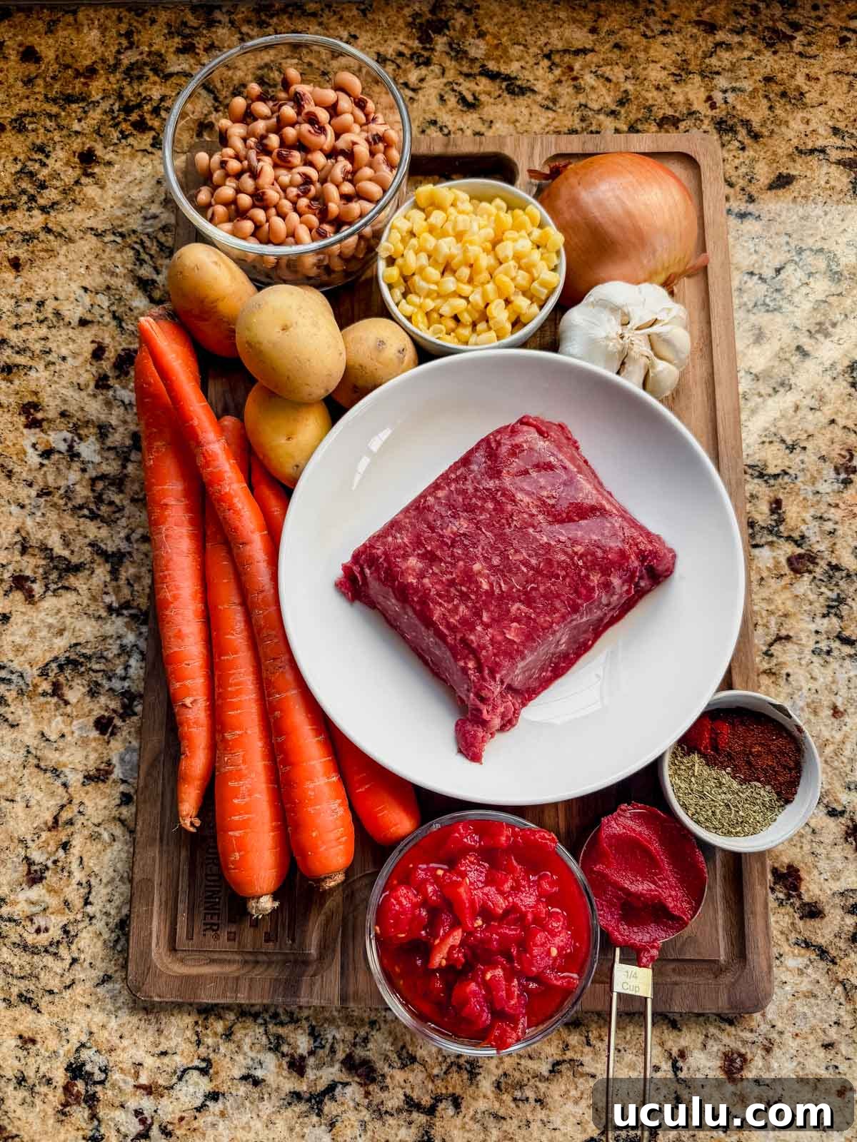 ground beef, carrots, corn, tomatoes potatoes, onion, and garlic on a cutting board.