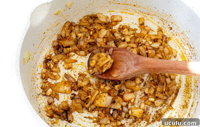 Onions and spices in a skillet with a wooden spoon stirring it.