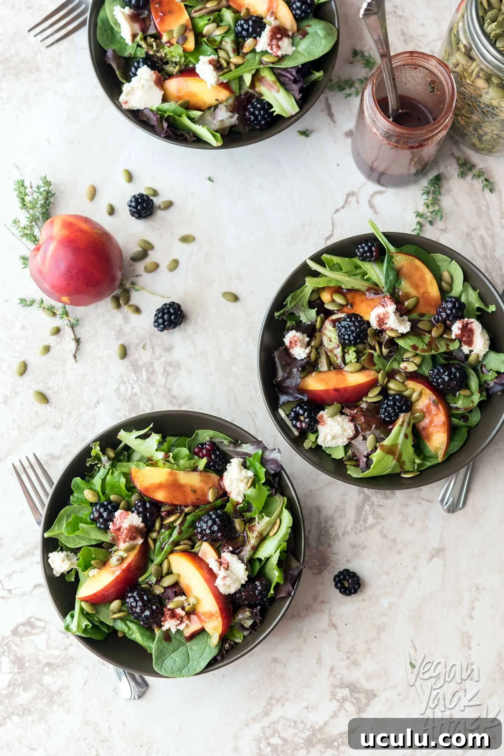 Three bowls of summery salad on a marble table top, next to seeds, a peach, and blackberries