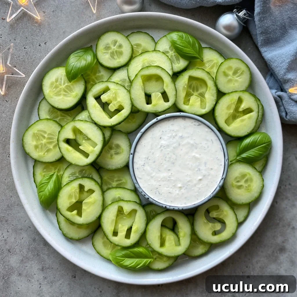 A festive Christmas Veggie Tray features cucumber slices with some spelling MERRY XMAS. Fresh basil leaves and a bowl of white dipping sauce sit at the center, surrounded by decorative lights and ornaments.