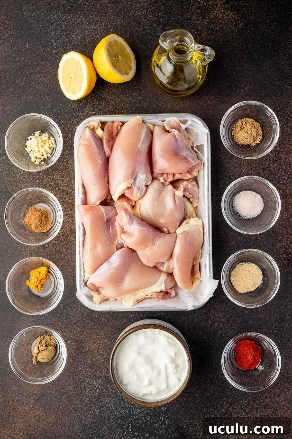 Raw chicken thighs in a tray surrounded by small bowls of spices, minced garlic, yogurt, lemon halves, and a bottle of olive oil on a dark surface.