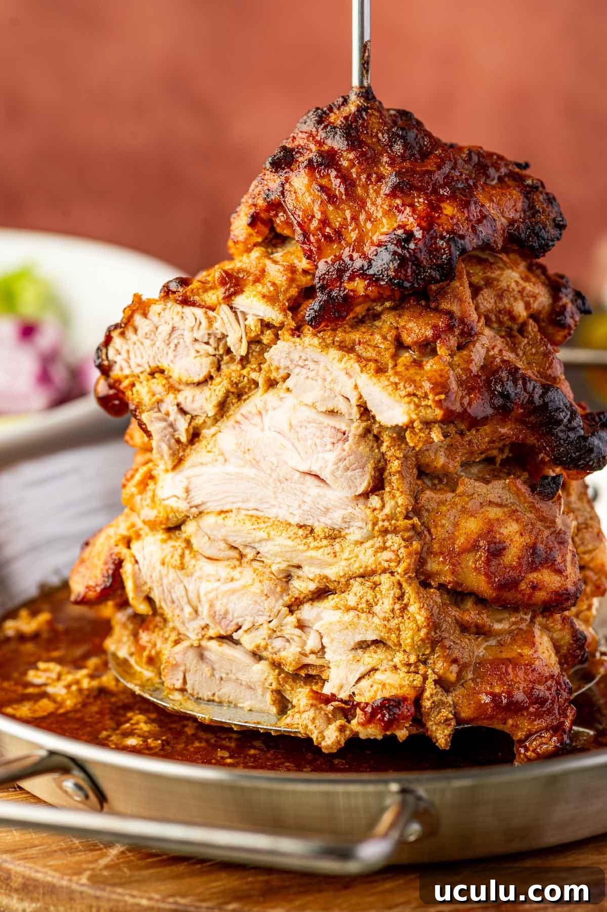 Stacked, marinated chicken shawarma roasting vertically on a spit in a metal pan, with some charred edges and juices collected at the bottom.
