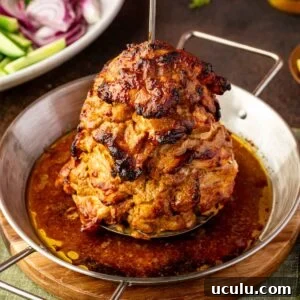 A stack of marinated and roasted chicken shawarma on a vertical spit in a round metal pan, with salad ingredients visible in the background.