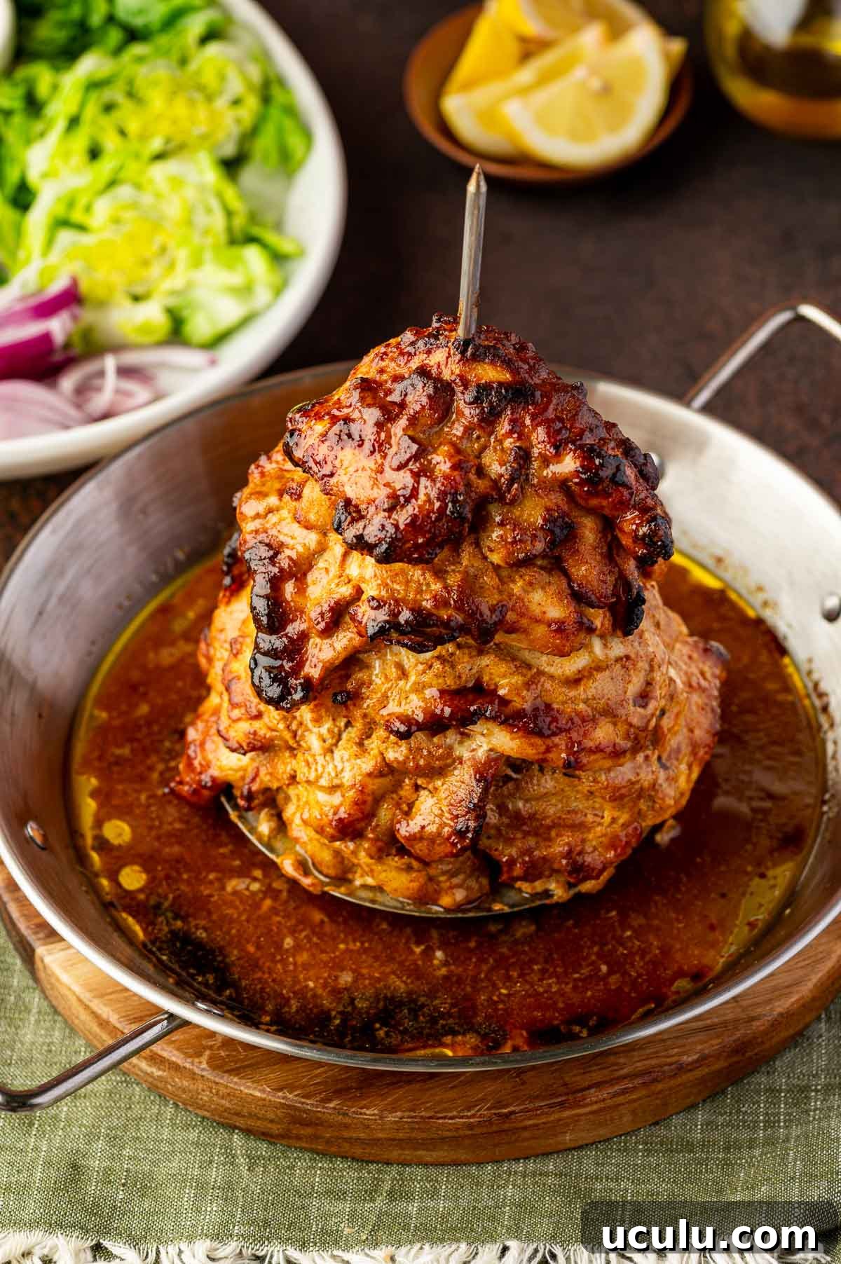 A stack of marinated chicken shawarma slices roasted on a vertical spit is served in a metal pan with juices, with salad and lemon wedges in the background.