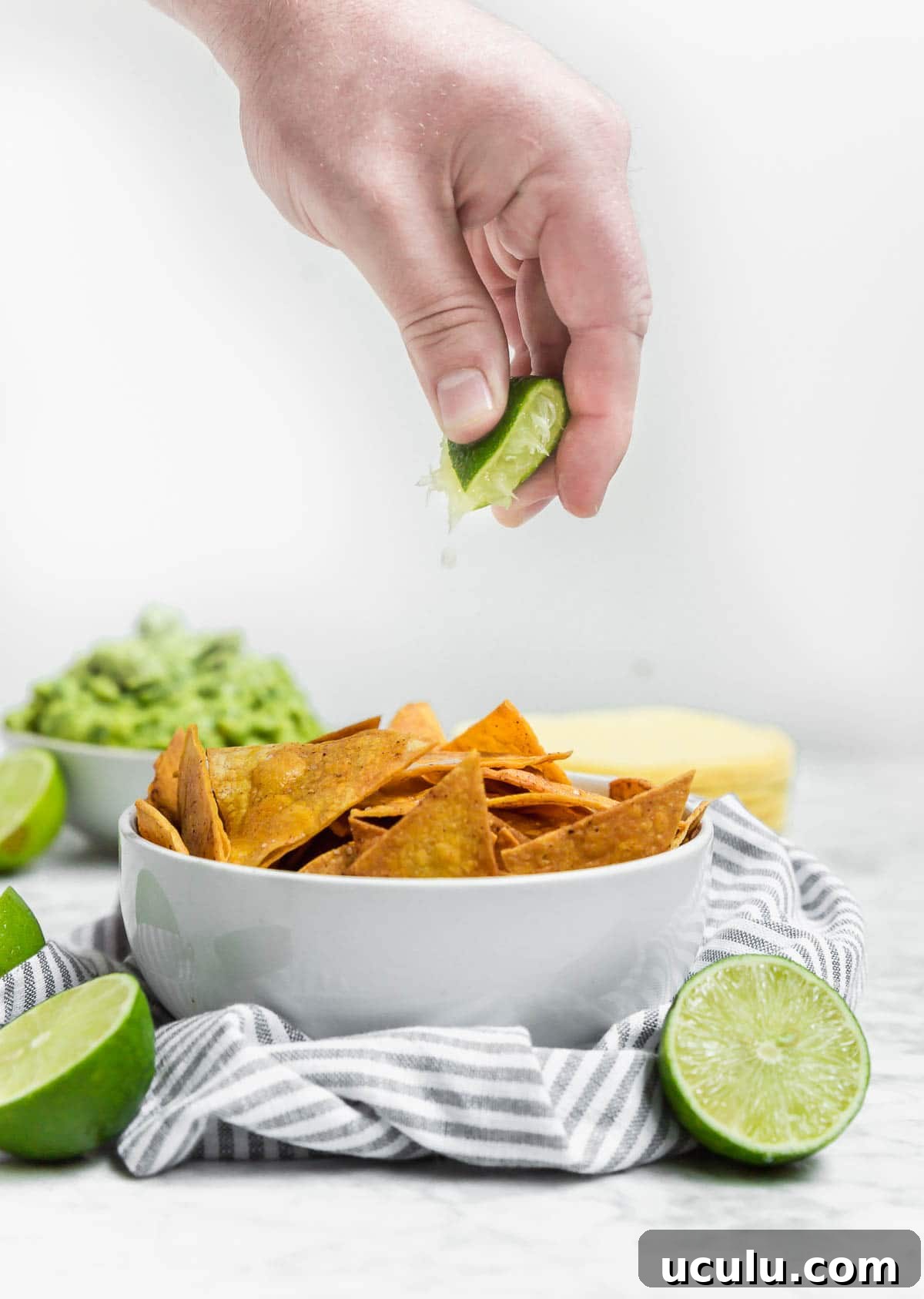 A hand squeezes a lime wedge over a bowl of Baked Chili Lime Tortilla Chips, with guacamole and more lime halves in the background, all set on a striped cloth.