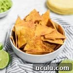 A white bowl filled with golden, crispy Baked Chili Lime Tortilla Chips sits on a striped cloth. Sliced limes and a stack of corn tortillas are nearby, with a bowl of guacamole in the background.