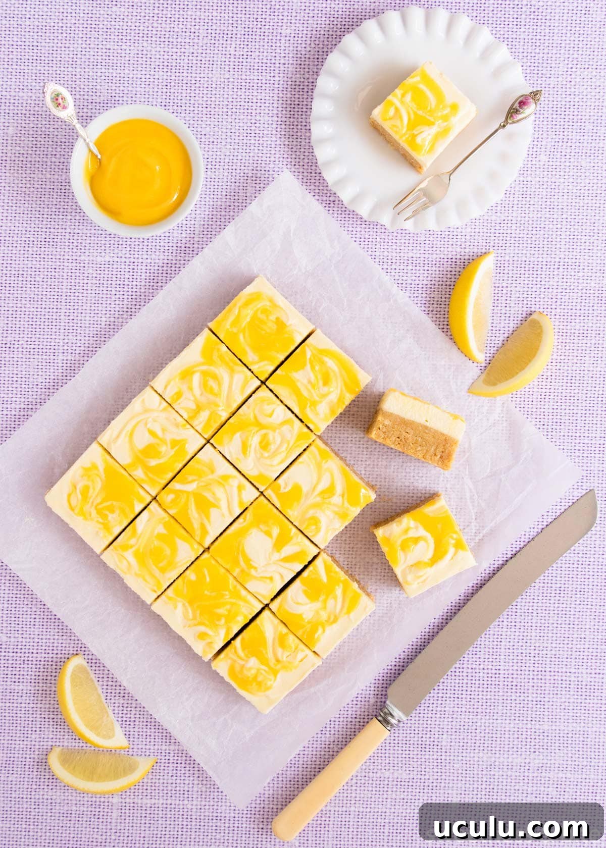 The slice, cut into squares on a pale purple background, with a knife and a bowl of lemon curd.