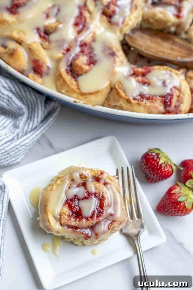 sourdough strawberry roll on a white plate with a fork on marble countertop. fresh strawberries are to the right and more rolls in a baking sheet are in the background
