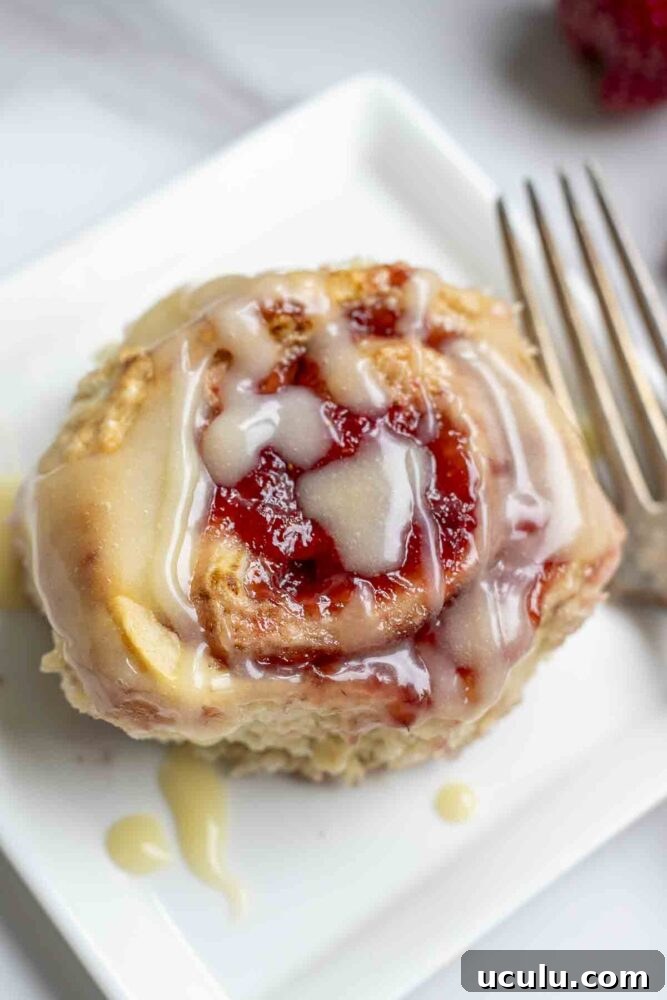 overhead photo of a sourdough strawberry sweet roll drizzled with icing on a white square plate with a antique fork to the right