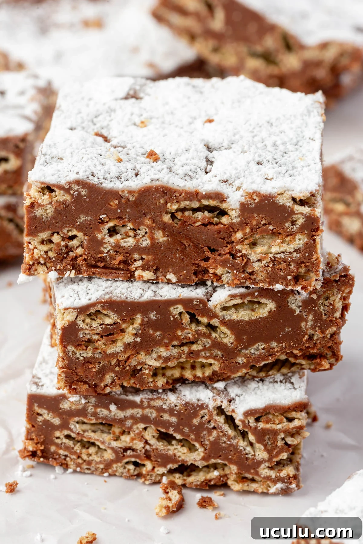 Close-up of three muddy buddy bars stacked on top of each other, showing a cross-section of Chex cereal coated in chocolate and dusted with powdered sugar.