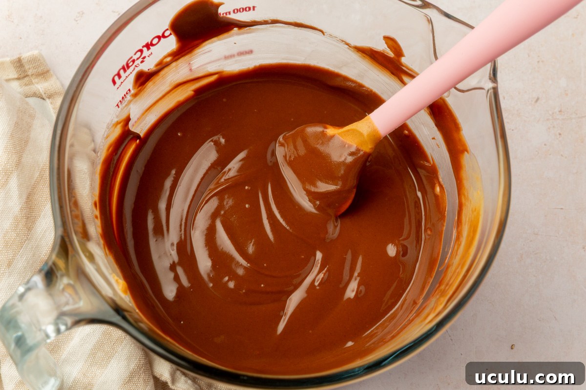 A glass bowl filled with smooth, glossy melted chocolate and peanut butter mixture, stirred with a pink spatula.