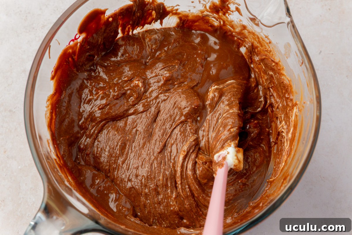 Melted chocolate, peanut butter, and marshmallow mixture in a glass bowl with a pink spatula mid-stir.