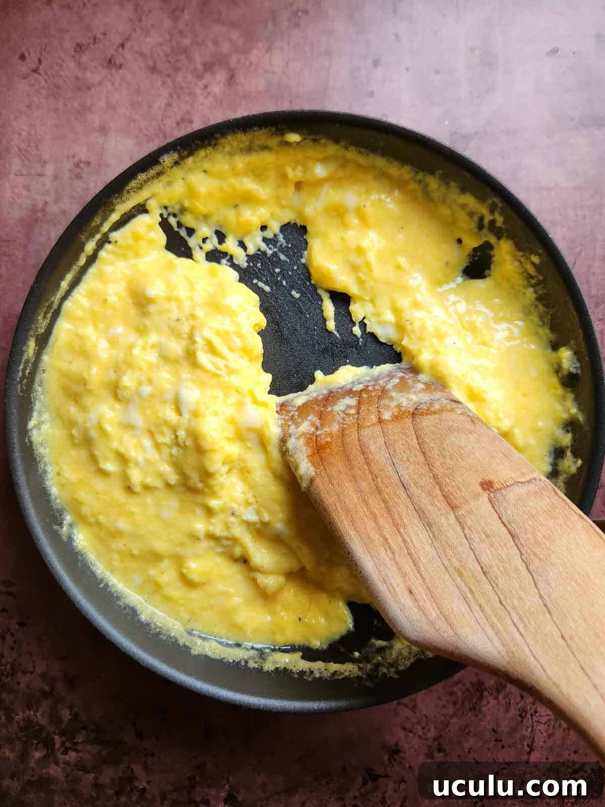 A wooden spoon shown moving scrambled eggs in a pan.