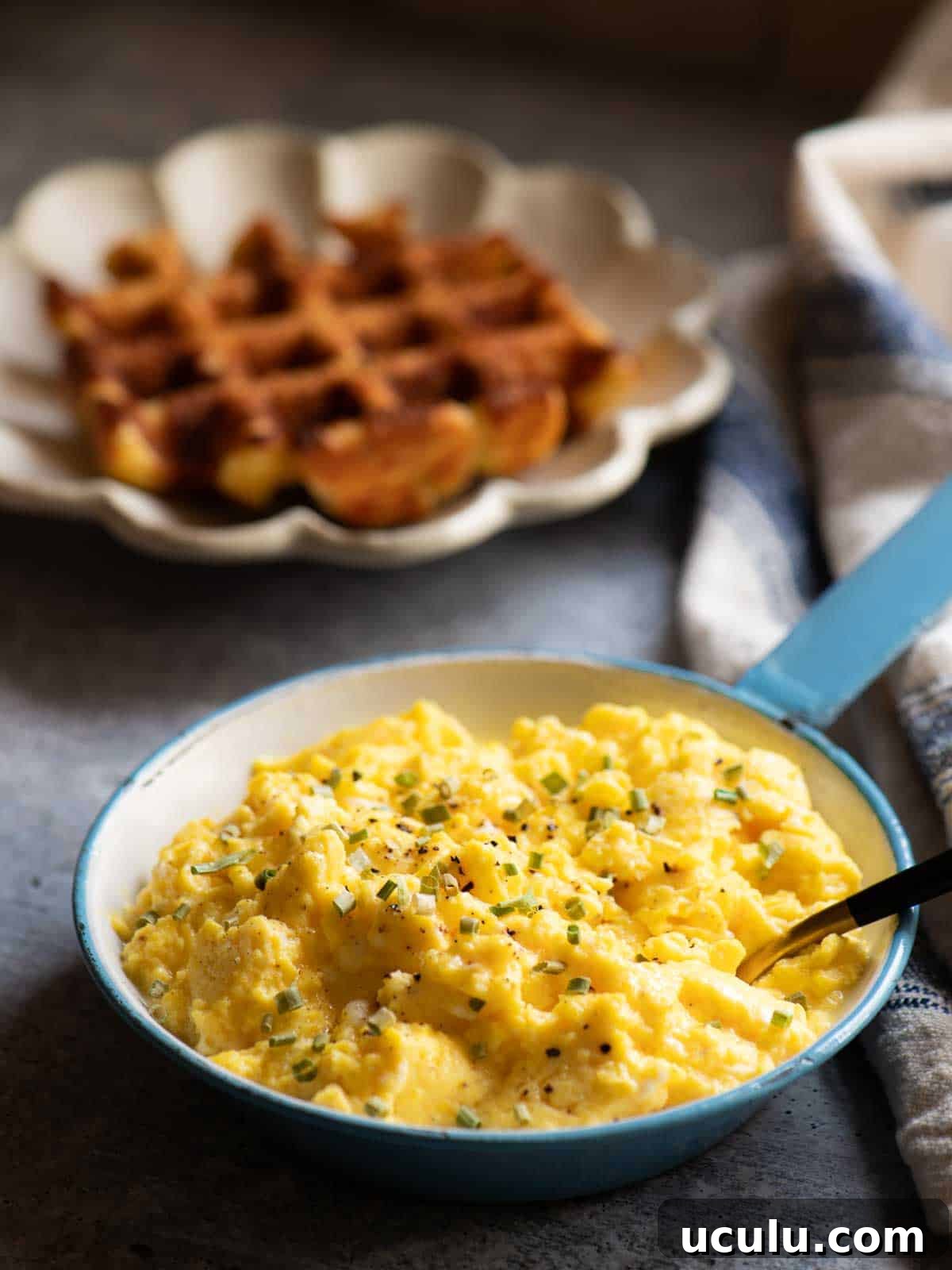 Scrambled eggs in a blue skillet with a waffle in the background.