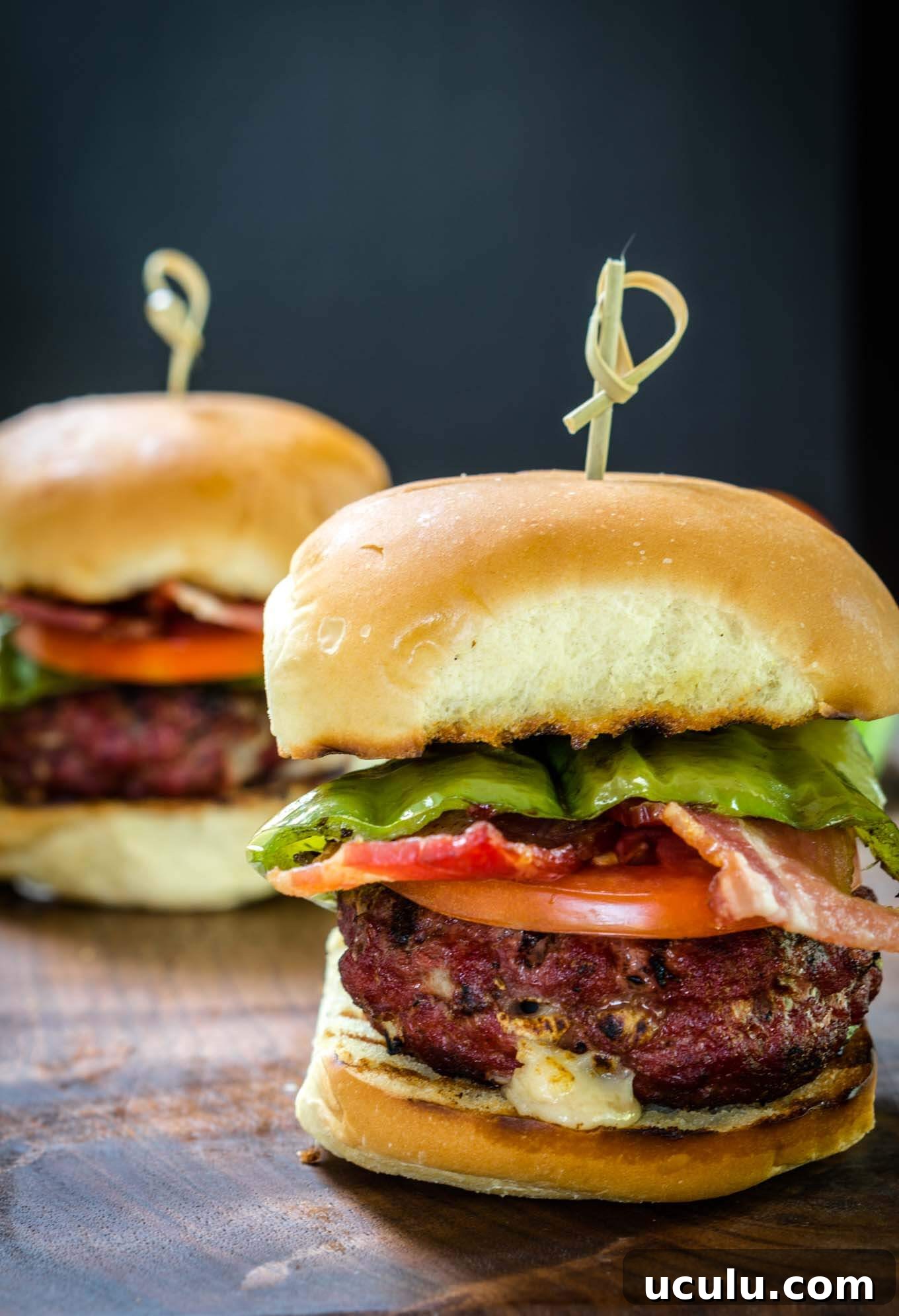 Sideview of a burger stuffed with hatch chile and Gruyere layered with lettuce, tomato, a hatch chile, and bacon secured with a bamboo skewer. A second burger sits in the background.
