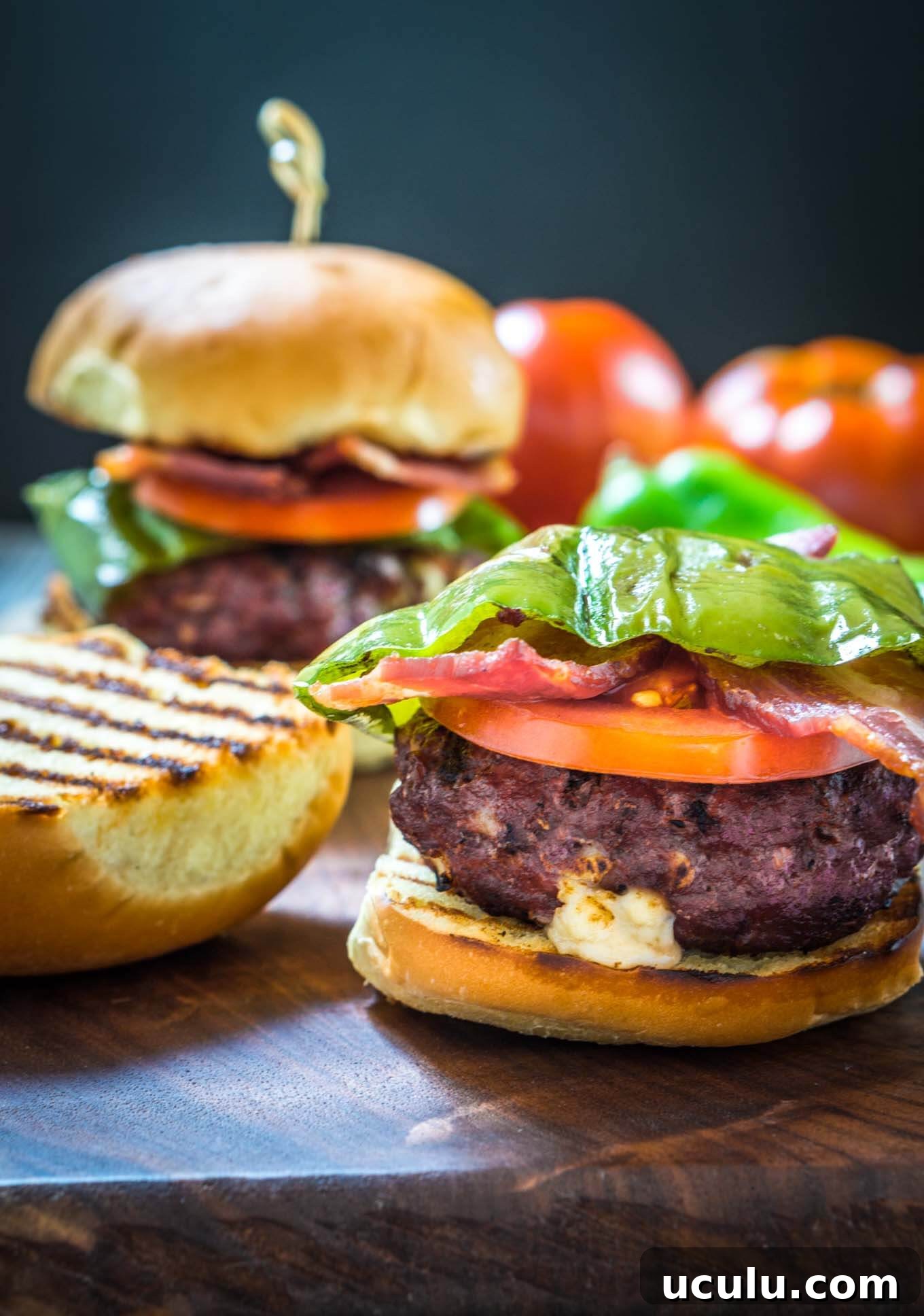 Sideview of a burger stuffed with hatch chile and Gruyere layered with lettuce, tomato, a hatch chile, and bacon with the top of the toasted bun sitting next to it. A second burger sits in the background next to fresh tomatoes.
