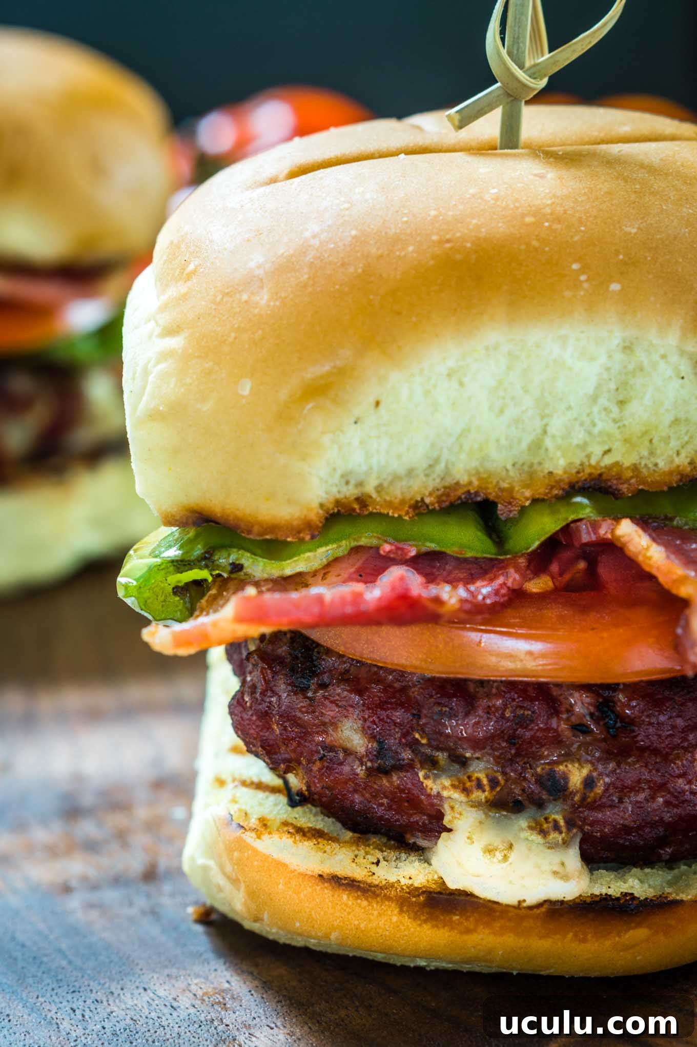 Sideview close-up of a burger stuffed with hatch chile and Gruyere layered with lettuce, tomato, a hatch chile, and bacon secured with a bamboo skewer. A second burger sits in the background.