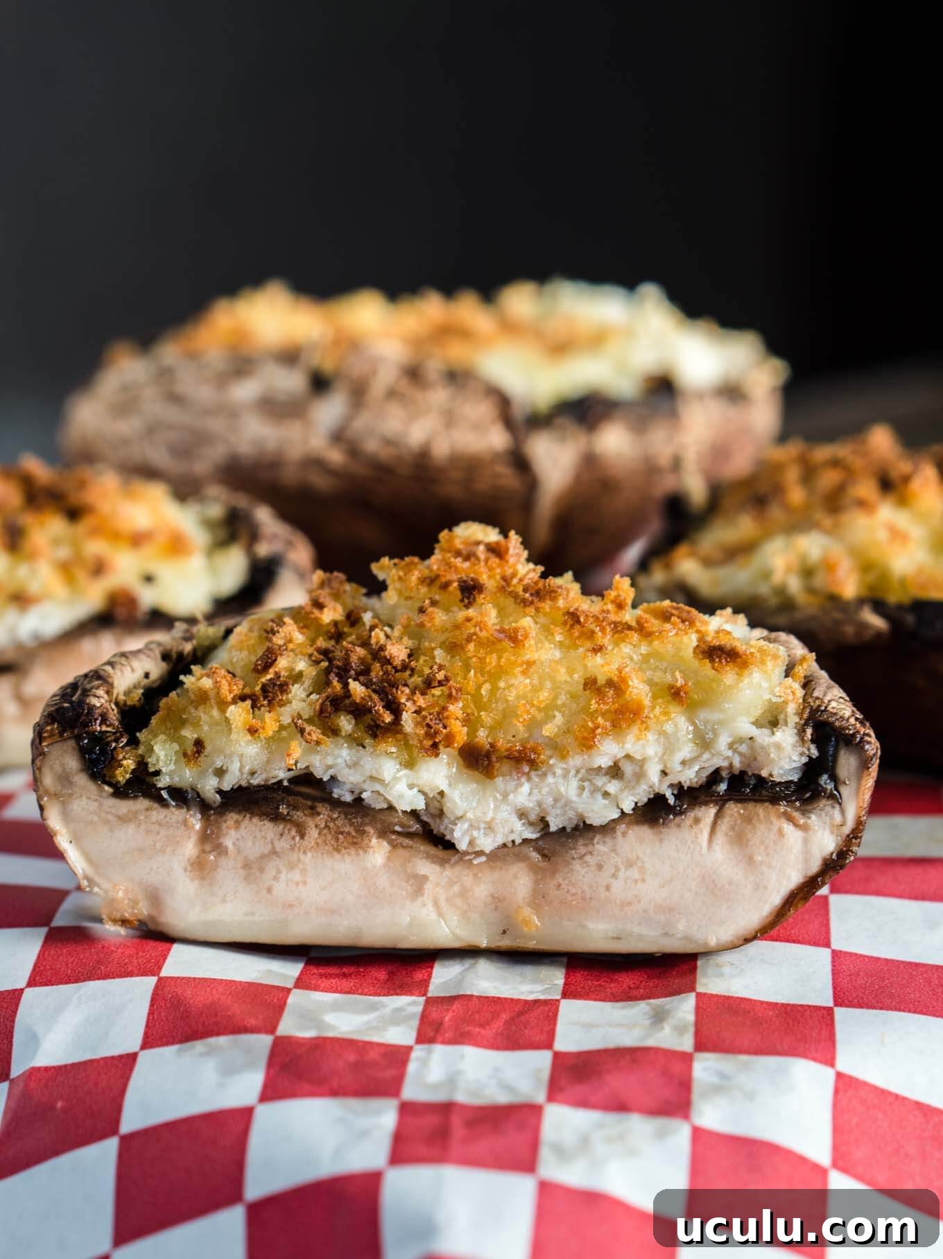 Crab stuffed portabello mushroom cut in half with more stuffed mushrooms sitting in the back over a red checked napkin.