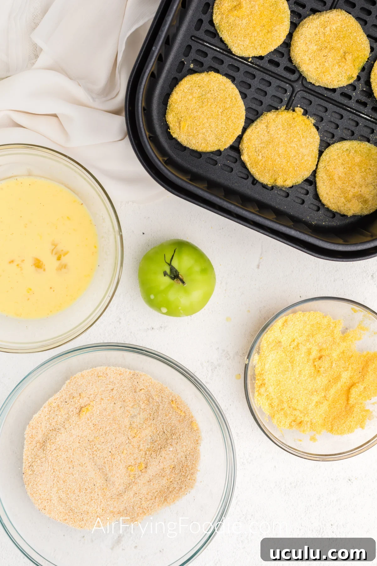 Coated green tomatoes in a single layer in the basket of the air fryer, surrounded by ingredients on a table.
